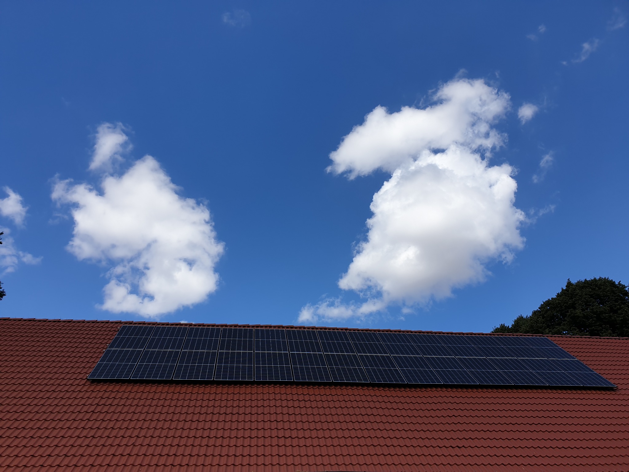 Panel słoneczny na dachu z czerwonej dachówki, na tle błękitnego nieba z białymi obłokami.