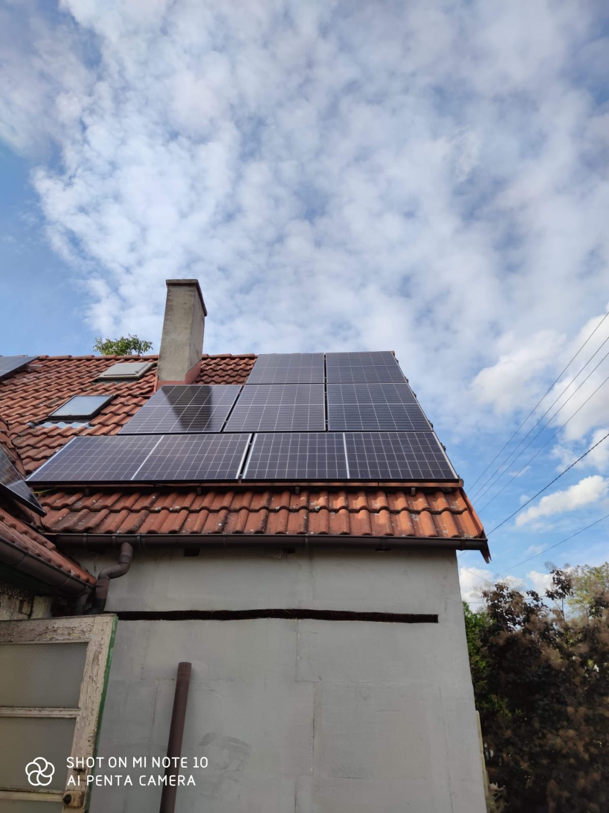 Panel słoneczny na starym, czerwonym dachu z kominem na tle błękitnego nieba z obłokami. Widoczna szara ściana budynku z oknem.