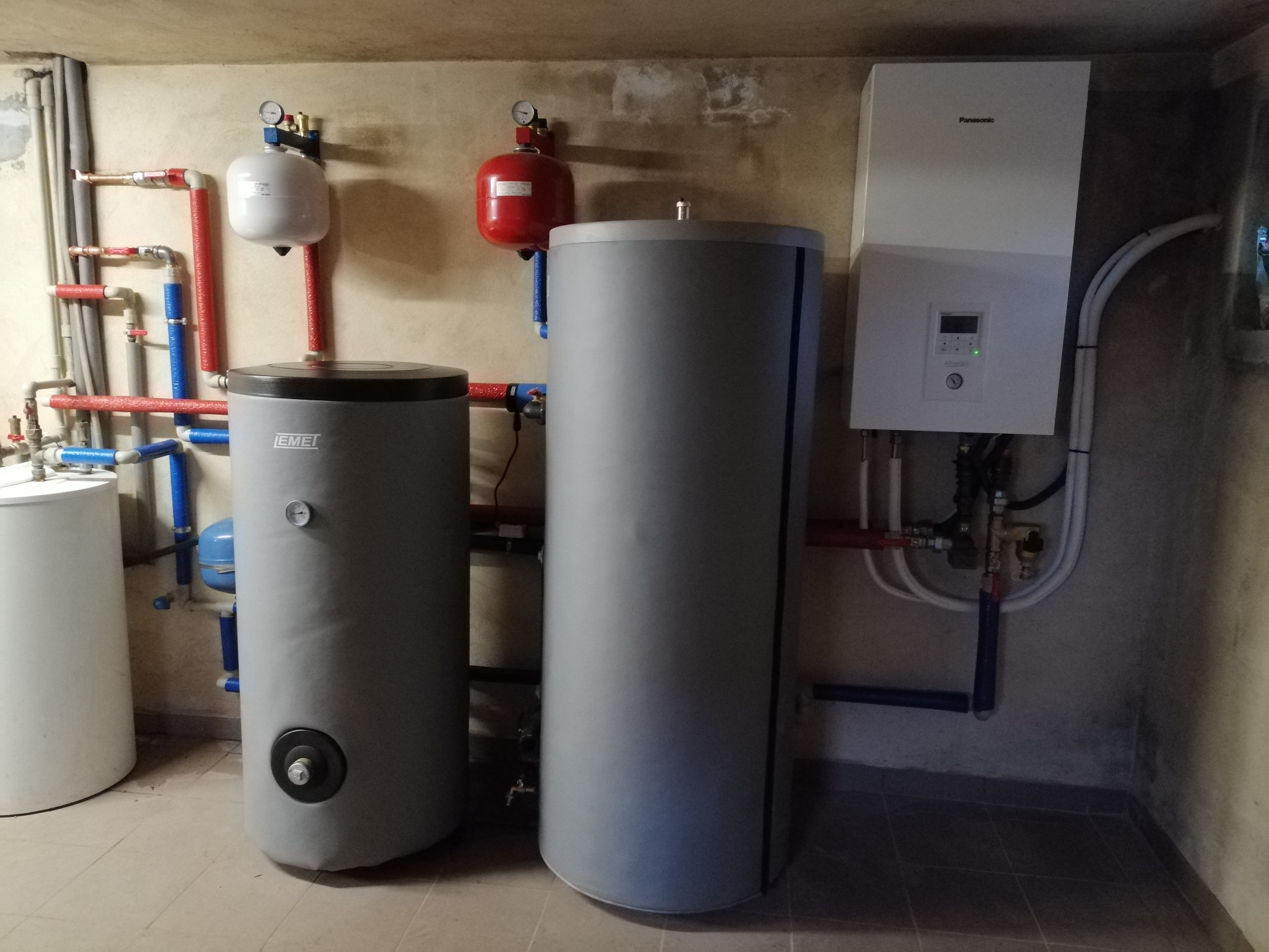 Instalacja centralnego ogrzewania z dwoma szarymi zbiornikami akumulacyjnymi, białym zbiornikiem wyrównawczym, czerwonym naczyniem przeponowym i białym modułem Panasonic z wyświetlaczem, połączona...