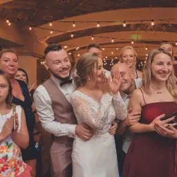 Uśmiechnięta para młoda w otoczeniu gości weselnych podczas występu artystycznego, oświetlona girlandami żar&oacute;wek.