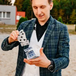 Uśmiechnięty iluzjonista w kraciastej marynarce i białej kamizelce prezentuje trik z unoszącymi się kartami na tle piaszczystej plaży i budynku.