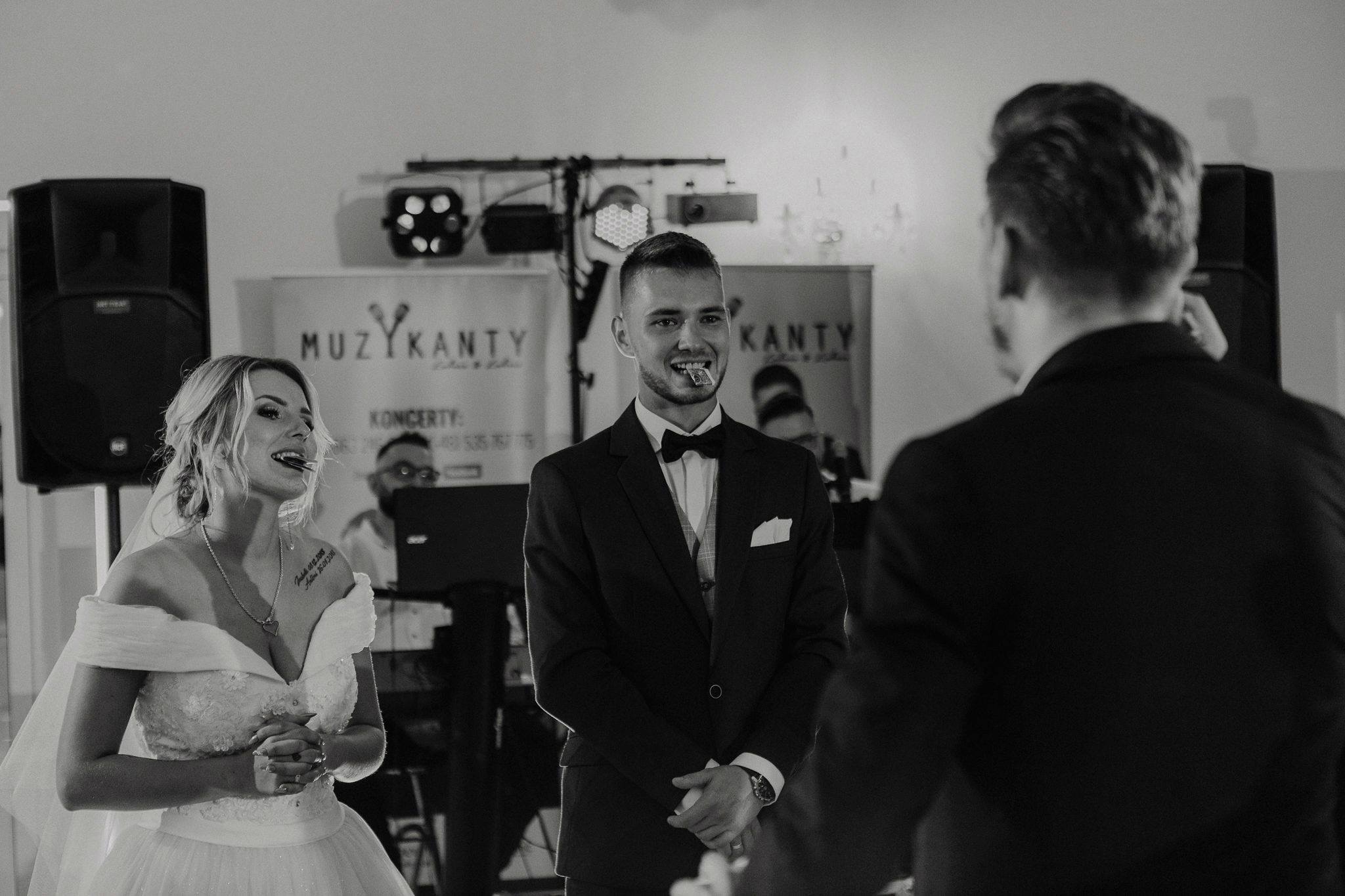 Czarno-białe zdjęcie uśmiechniętego mężczyzny w garniturze z kartą w ustach, stojącego na tle sceny z parą młodą i sprzętem nagłośnieniowym.