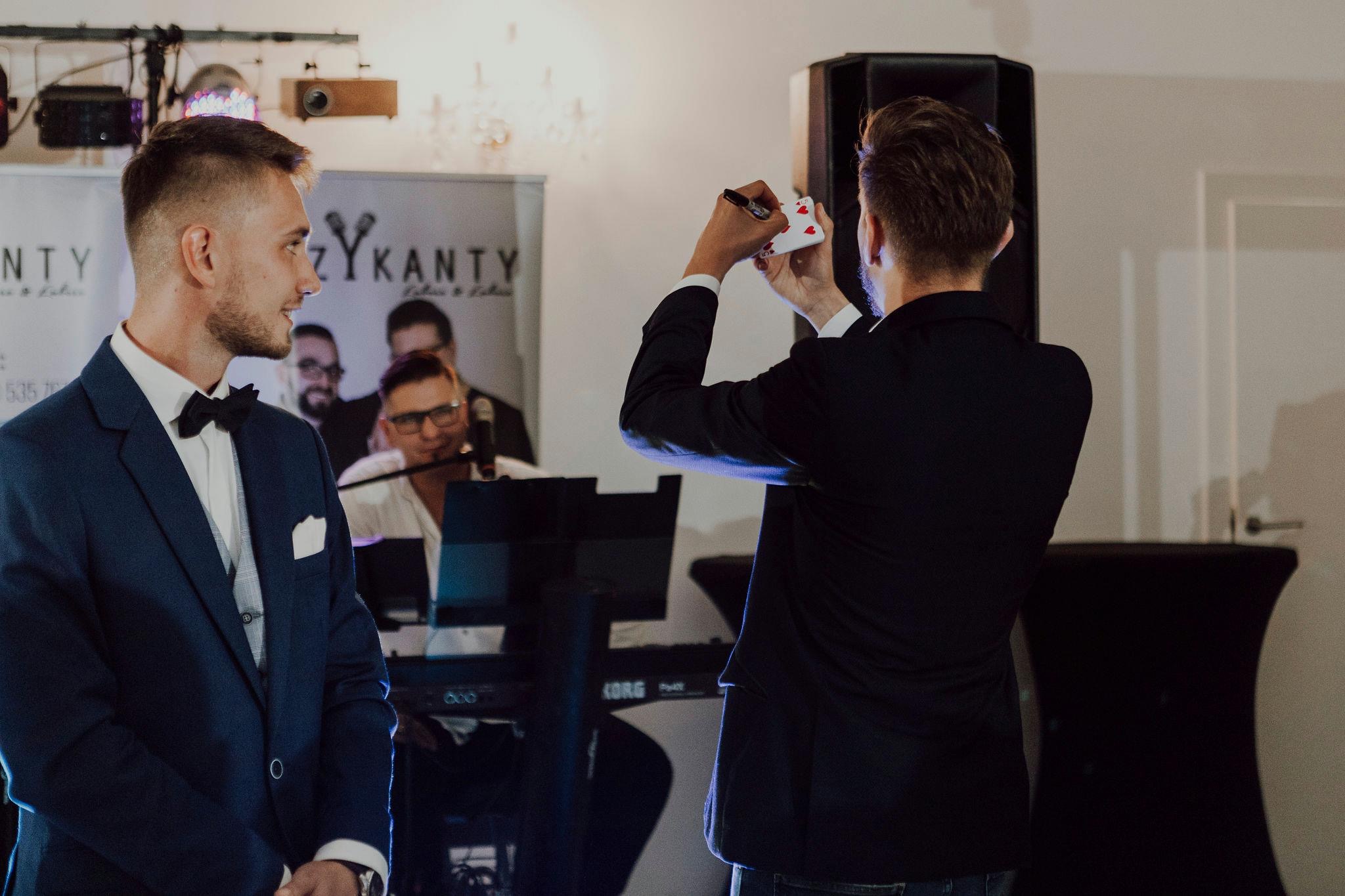 Iluzjonista w ciemnym garniturze pisze markerem na karcie z czerwonymi sercami, drugi mężczyzna w niebieskim garniturze z muchą patrzy z zaciekawieniem, w tle zespół muzyczny z logotypem firmy.