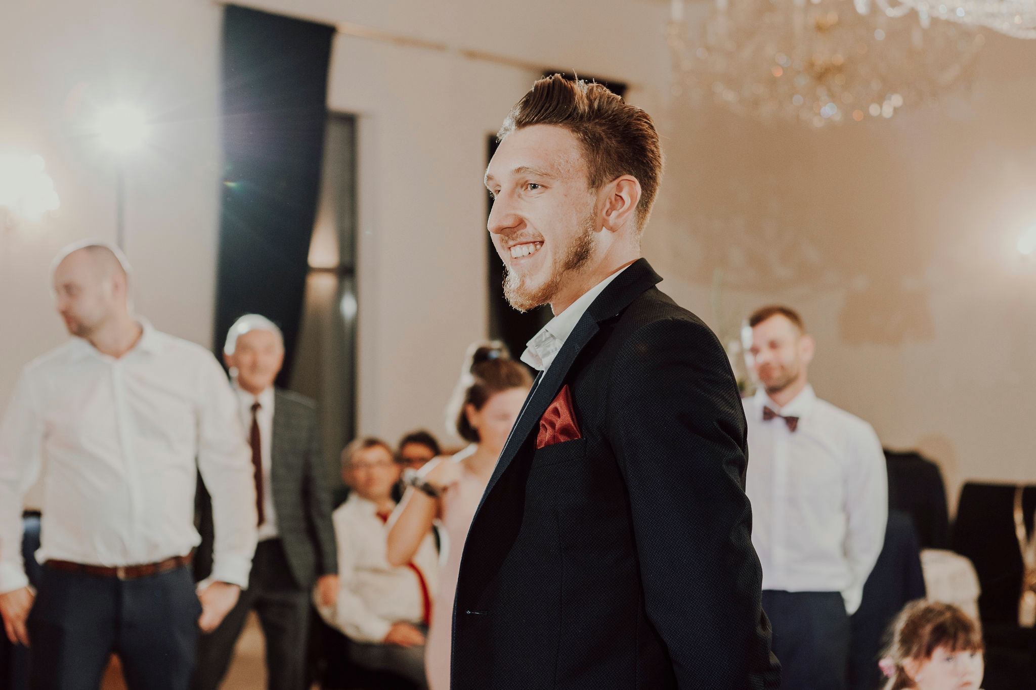 Uśmiechnięty mężczyzna w ciemnej marynarce z czerwoną poszetką, stojący na tle sali z gośćmi i żyrandolem, podczas imprezy.