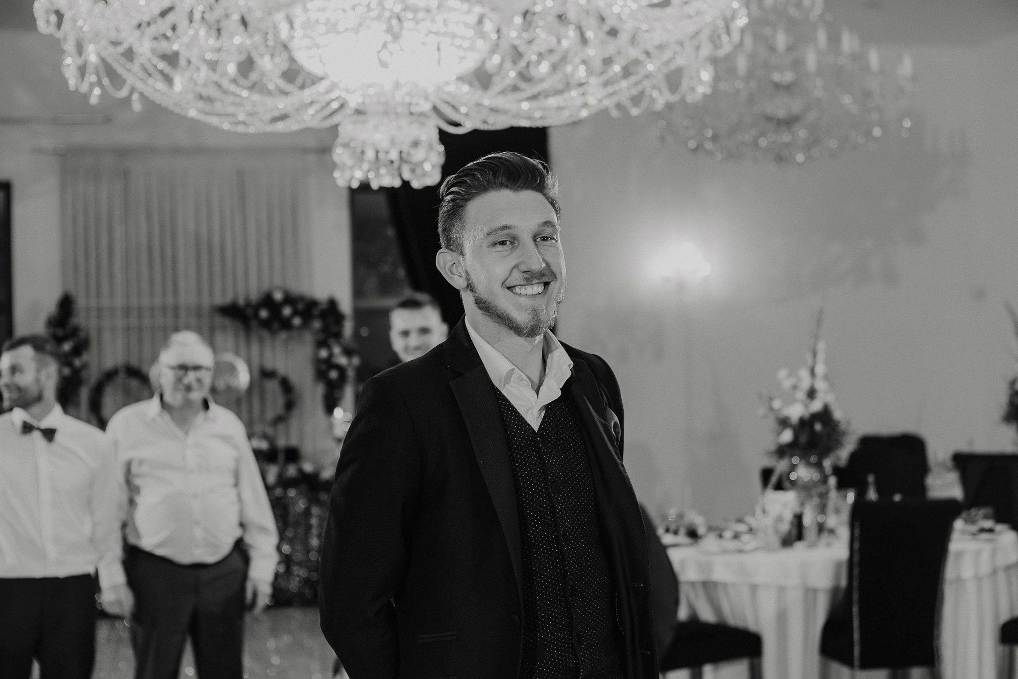 Uśmiechnięty mężczyzna w garniturze na tle sali bankietowej z kryształowym żyrandolem.