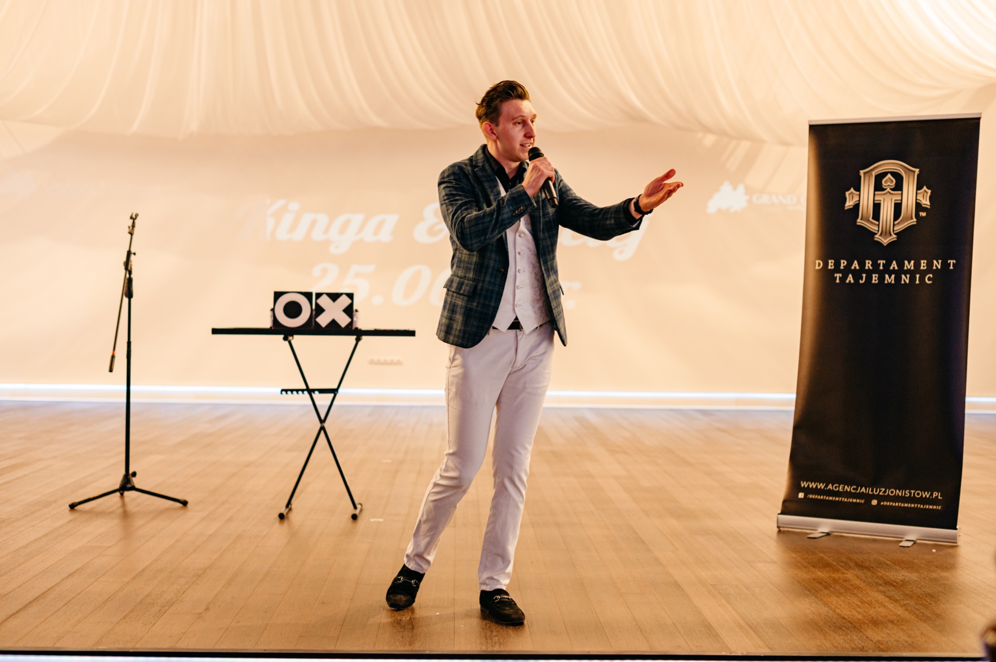 Mężczyzna w kraciastej marynarce i białej kamizelce, trzymający mikrofon i gestykulujący na scenie podczas występu, obok stolik z rekwizytami i roll-up z logo agencji.