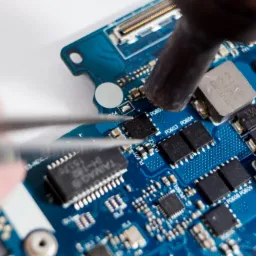 Detail of a blue circuit board being worked on with tweezers and a soldering iron, showing various electronic components and their labels.