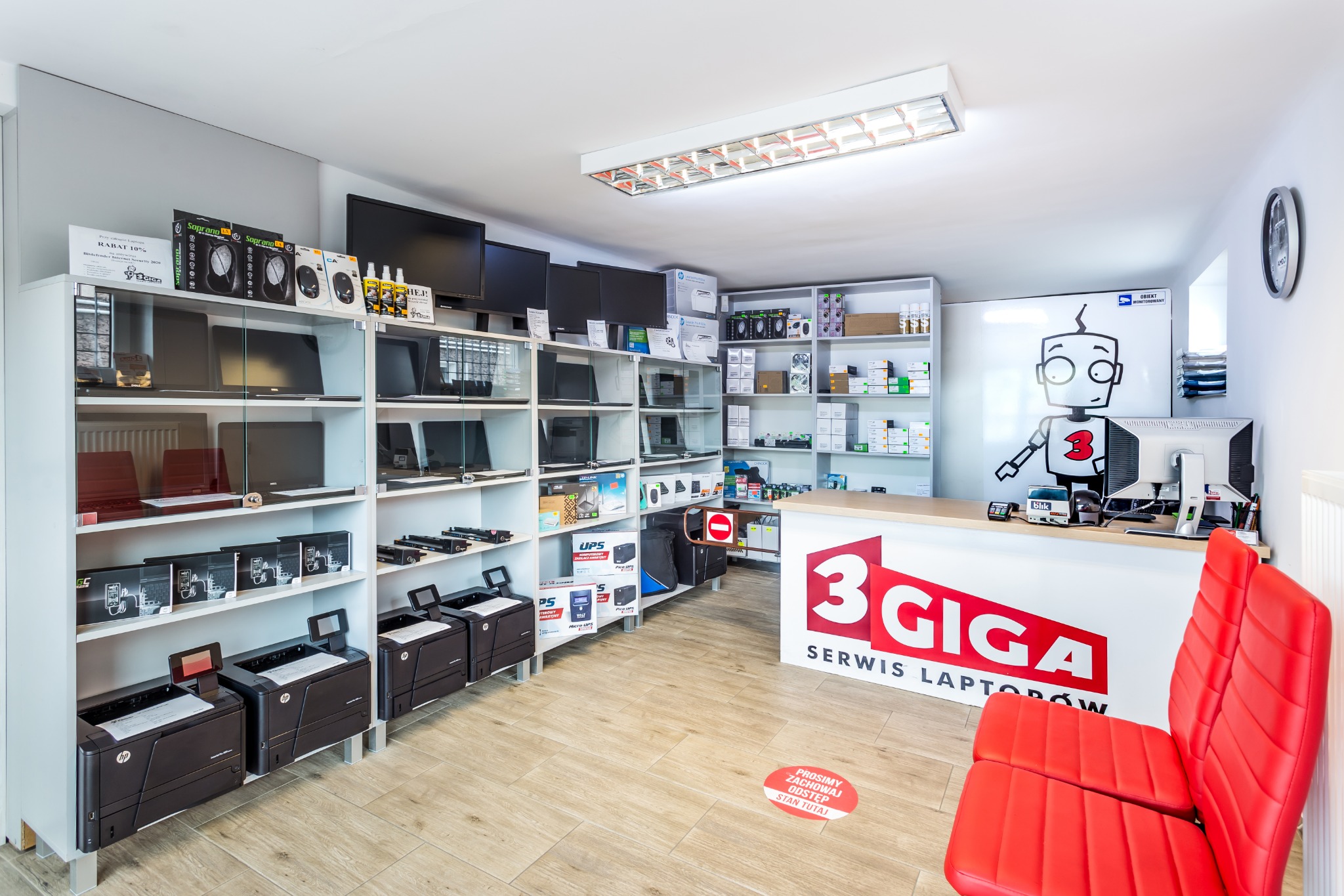 Wnętrze punktu serwisowego z ladą, regałami pełnymi laptopów, monitorów i drukarek, oraz naklejką na podłodze informującą o zachowaniu odstępu.