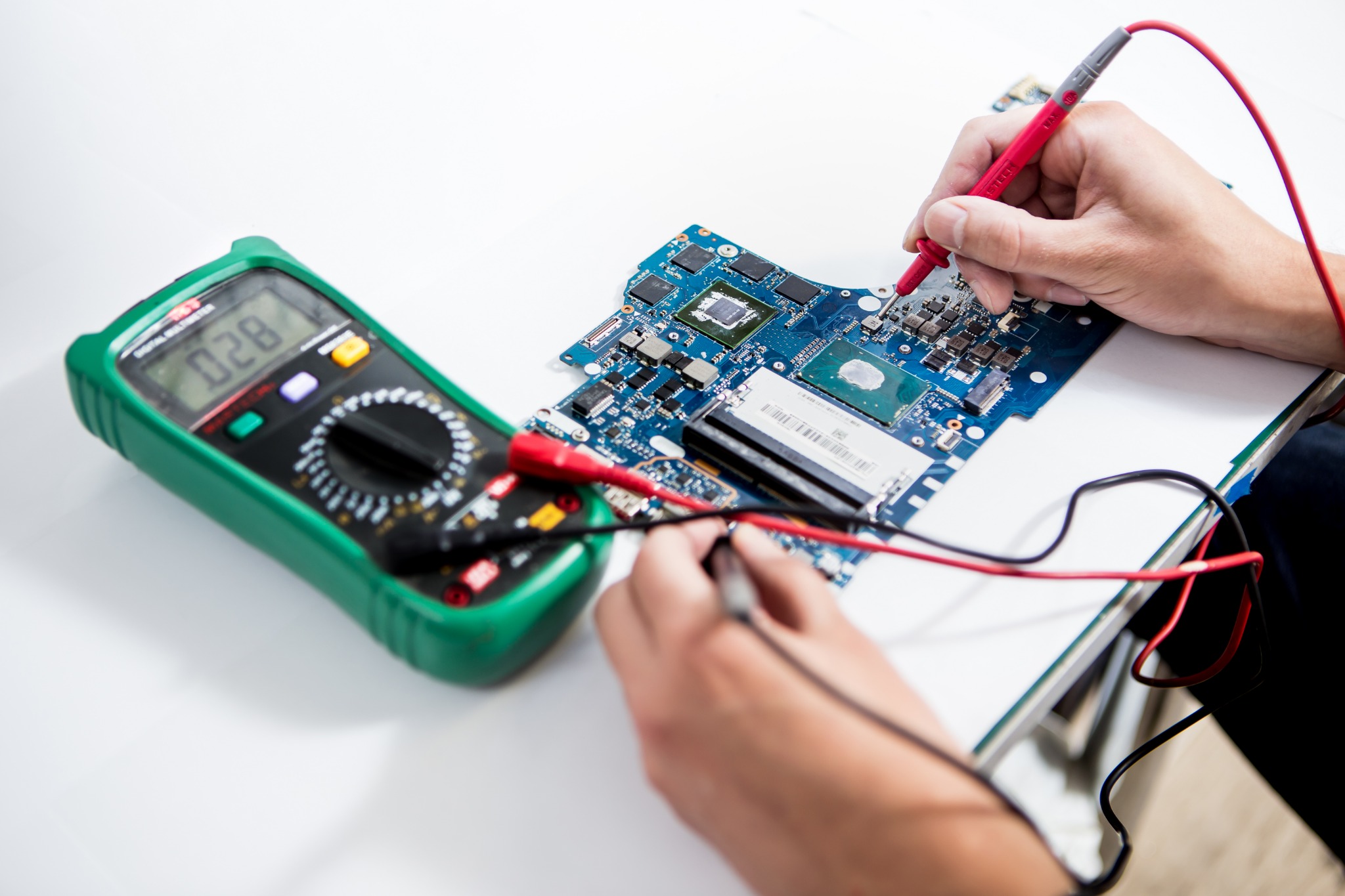 Elektronik mierzy napięcie na płycie głównej laptopa za pomocą multimetru z zieloną obudową, wyświetlającego wartość 0.28.
