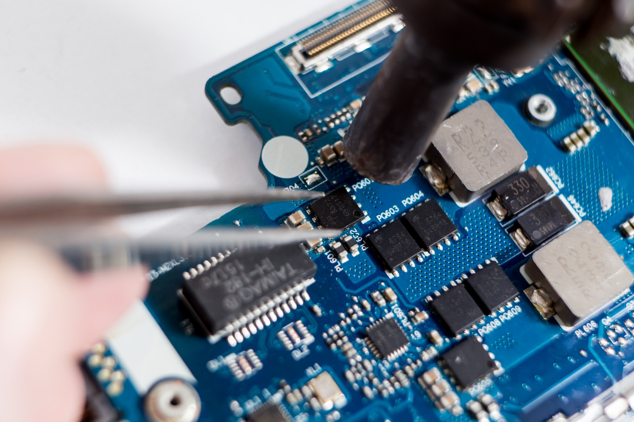 Detail of a blue circuit board being worked on with tweezers and a soldering iron, showing various electronic components and their labels.