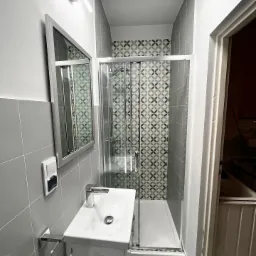 Nowoczesna łazienka z szarymi płytkami, kabiną prysznicową z wzorzystą ścianą i białą umywalką z lustrem, oświetlona podłużną lampą LED.