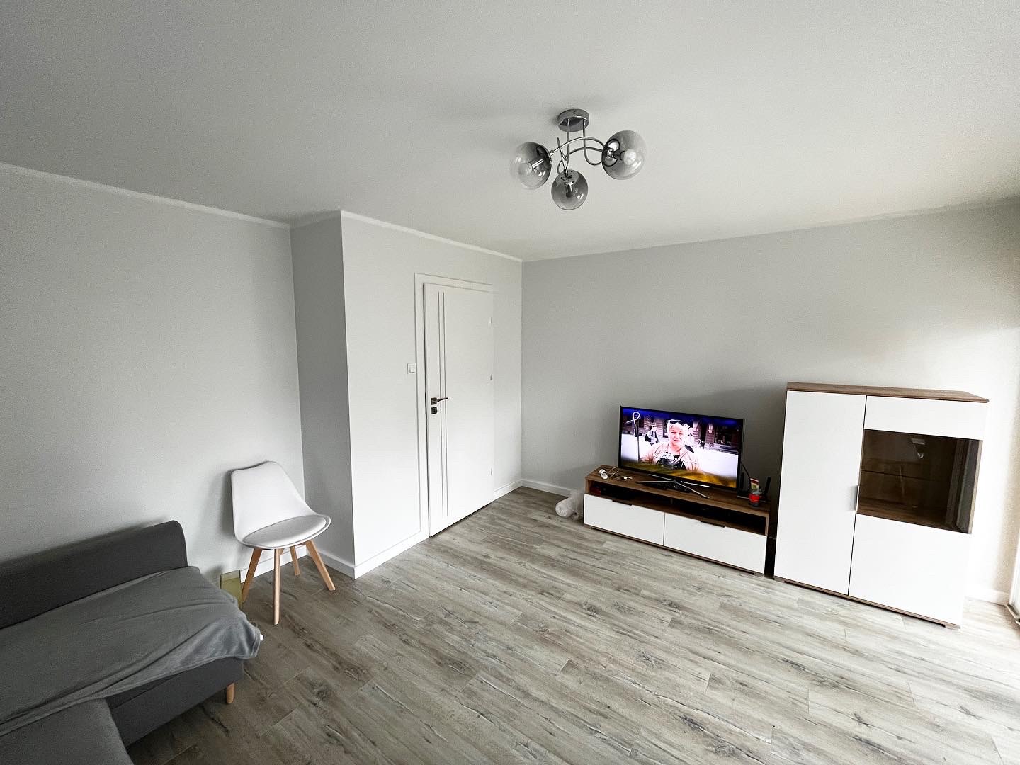 Jasne wnętrze pokoju po remoncie, z szarymi ścianami, panelami podłogowymi w odcieniach szarości, nowoczesną lampą sufitową z czterema szklanymi kloszami, telewizorem, białą komodą i szafką...