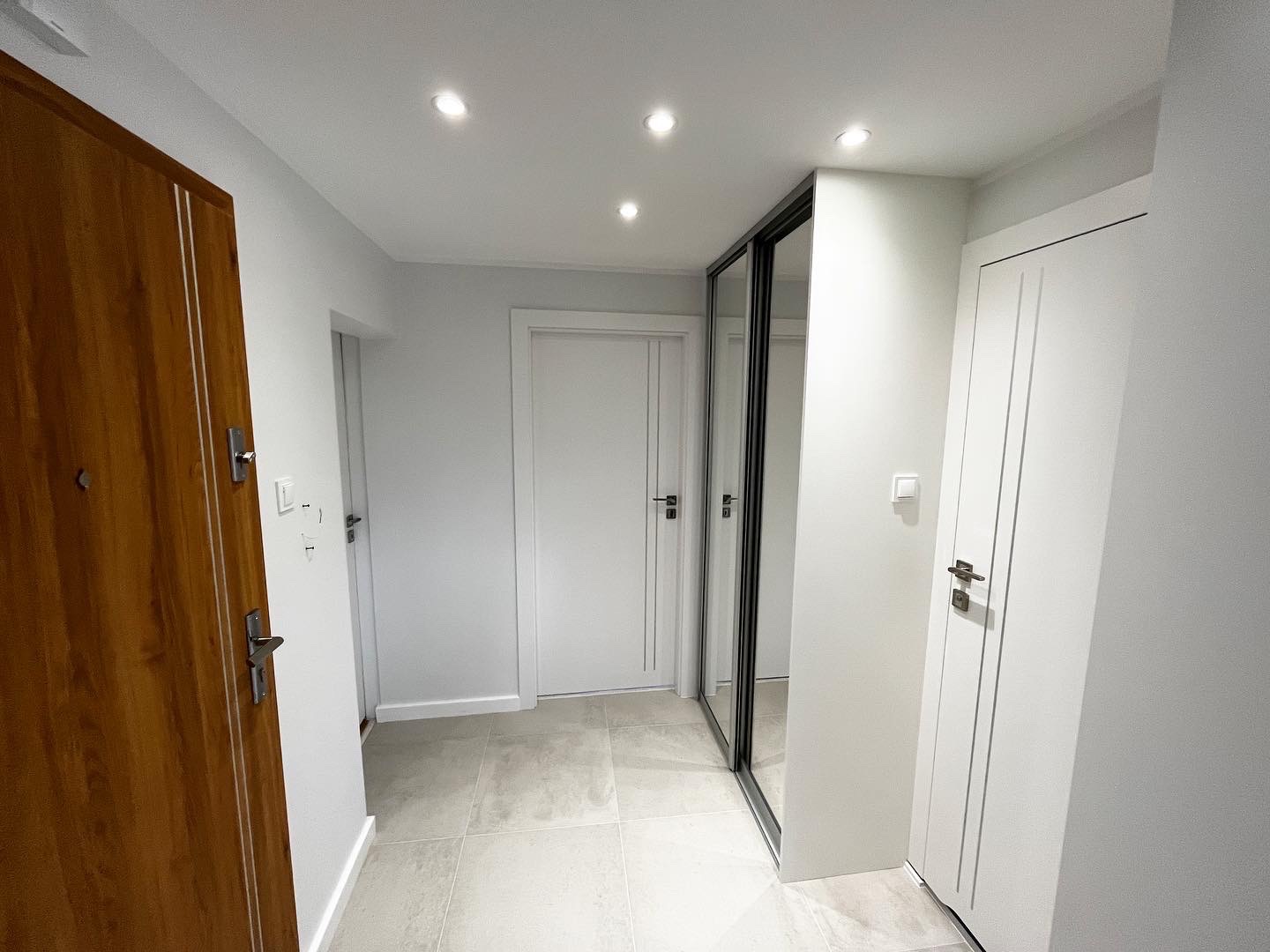 Nowoczesny przedpokój po remoncie: drewniane drzwi wejściowe, białe drzwi wewnętrzne z frezowanymi liniami, lustrzana szafa przesuwna w czarnej ramie i szara płytka podłogowa.