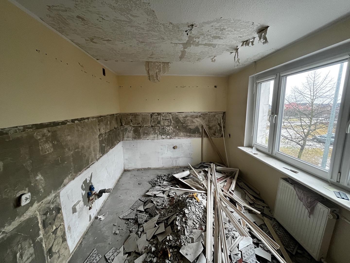 Pomieszczenie w trakcie demontażu płytek i przygotowania ścian pod remont; widoczne fragmenty starej instalacji, resztki płytek na podłodze i uszkodzony sufit z odpadającą farbą.