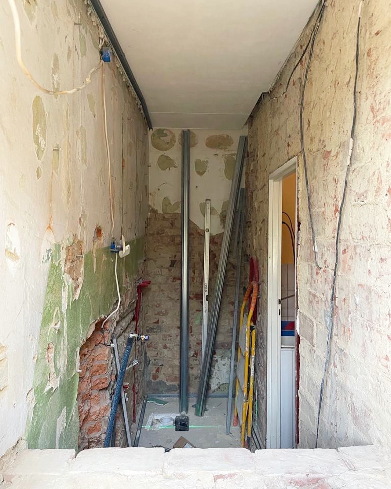 Wnętrze w trakcie renowacji: odsłonięta cegła, fragmenty starej farby, instalacje, profile metalowe i drabina oparta o framugę drzwi do łazienki. Widoczne ślady po kuciu ścian i przygotowania...