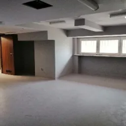 Wnętrze surowego pomieszczenia w Bardo po wylewce, widoczne okna z kratami i częściowo otwarte drzwi.