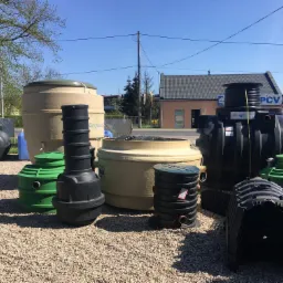 Ekspozycja różnych typów szamb betonowych i przydomowych oczyszczalni ścieków na żwirowym podłożu, widoczne różne kolory (czarny, beżowy, zielony) i konstrukcje, w tle budynek z szyldem PCV.