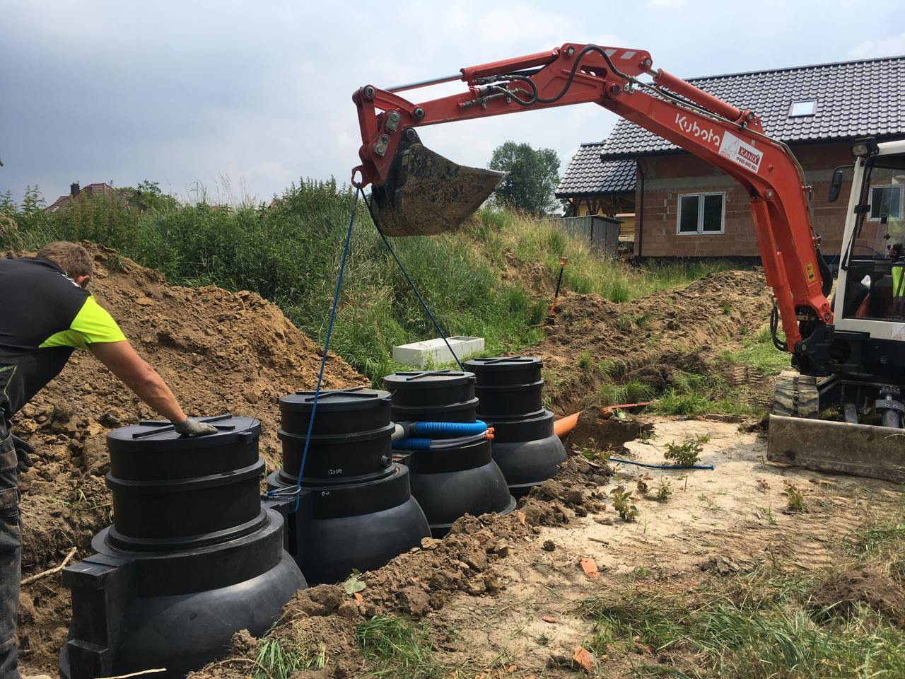 Montaż modułowej przydomowej oczyszczalni ścieków z użyciem koparki Kubota, widoczny pracownik w odzieży roboczej.