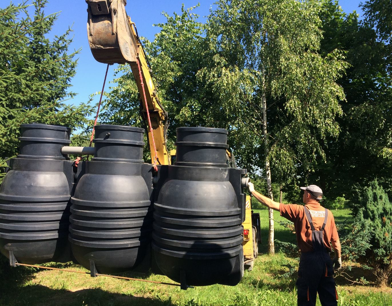 Żuraw przenosi czarny, zbiornik segmentowy na tle zieleni, pracownik w kombinezonie nadzoruje proces.