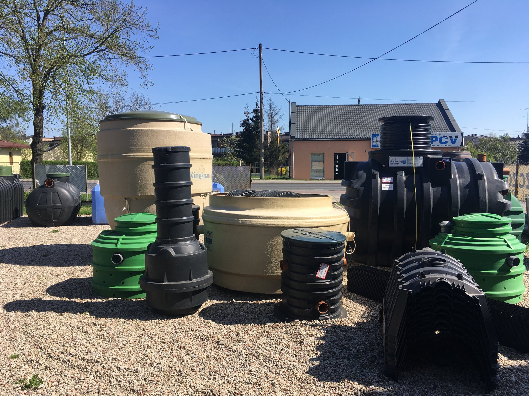 Ekspozycja różnych typów szamb betonowych i przydomowych oczyszczalni ścieków na żwirowym podłożu, widoczne różne kolory (czarny, beżowy, zielony) i konstrukcje, w tle budynek z szyldem PCV.