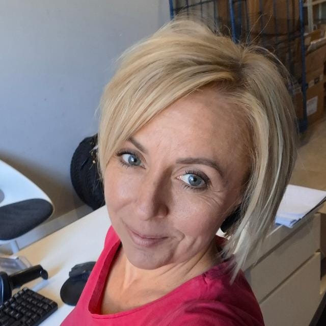 Portret kobiety z krótkimi blond włosami, uśmiechającej się do kamery, w biurowym otoczeniu z biurkiem i komputerem. Widoczne regały w tle.