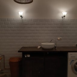 ELCON Electris Adam Łoś - Umywalka nablatowa na ciemnym blacie, nad nią dwa kinkiety w kształcie kwiatów i pleciony abażur, w tle białe płytki imitujące cegłę, obok pralka i kosz na pranie.