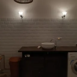 Umywalka nablatowa na ciemnym blacie, nad nią dwa kinkiety w kształcie kwiatów i pleciony abażur, w tle białe płytki imitujące cegłę, obok pralka i kosz na pranie.