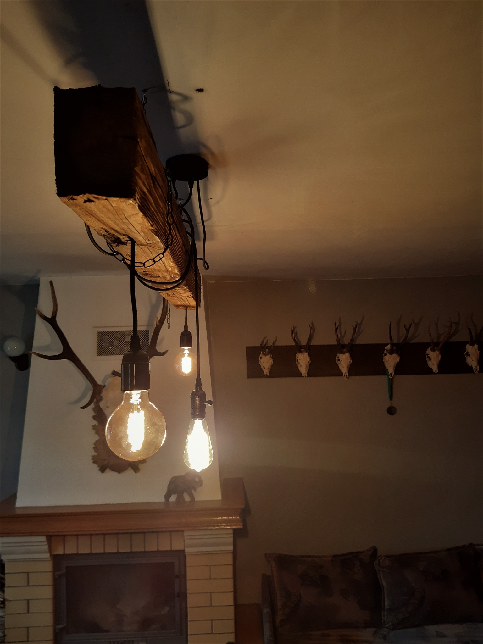 Rustykalna lampa wisząca z drewnianej belki i żarówek Edisona, zawieszona na łańcuchach, w tle kominek z dekoracjami myśliwskimi i kolekcja poroży na ścianie.