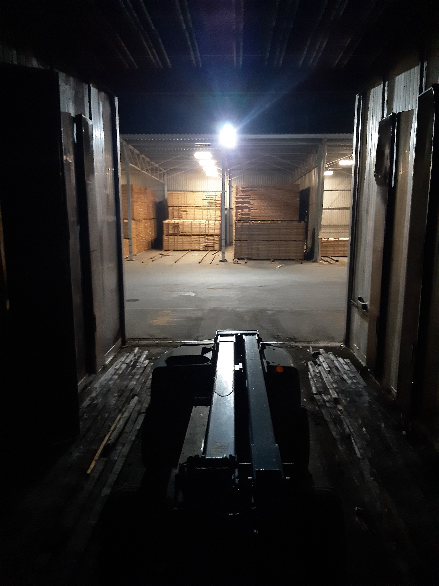 Wnętrze ciemnego kontenera z widokiem na magazyn desek oświetlony lampami, widziany z perspektywy operatora wózka widłowego.