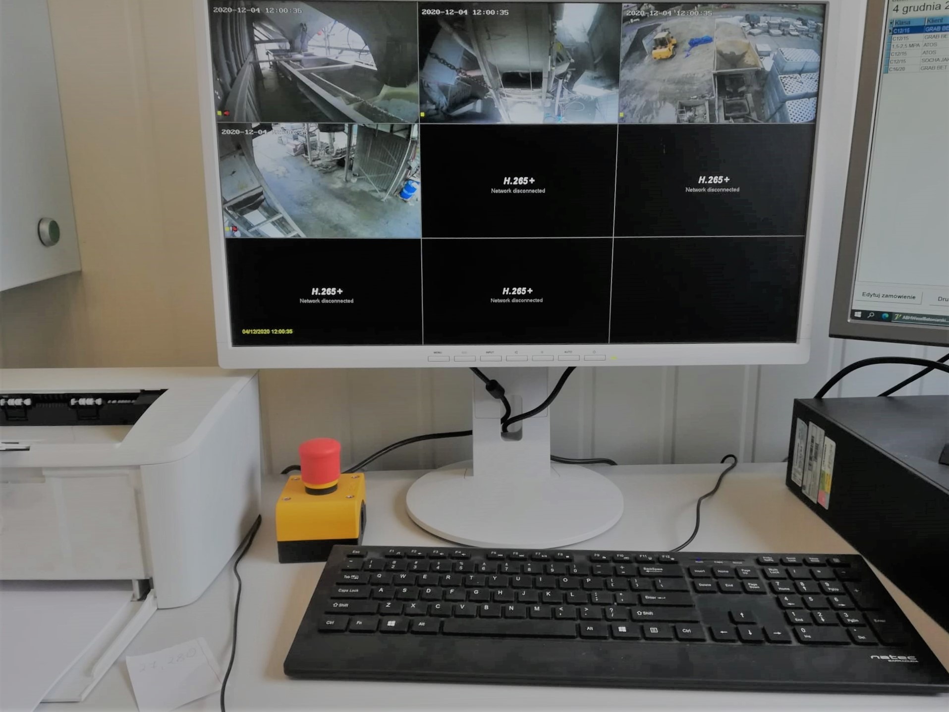 Monitor wyświetlający obraz z kamer przemysłowych monitorujących proces produkcji betonu, obok klawiatura, drukarka i przycisk awaryjny.