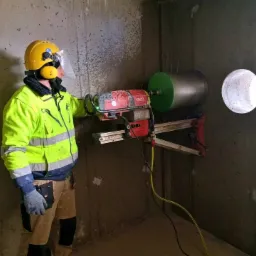 Pracownik w kasku i odblaskowej kurtce wierci otwór w betonowej ścianie za pomocą wiertnicy Hilti z zielonym wiertłem koronowym.
