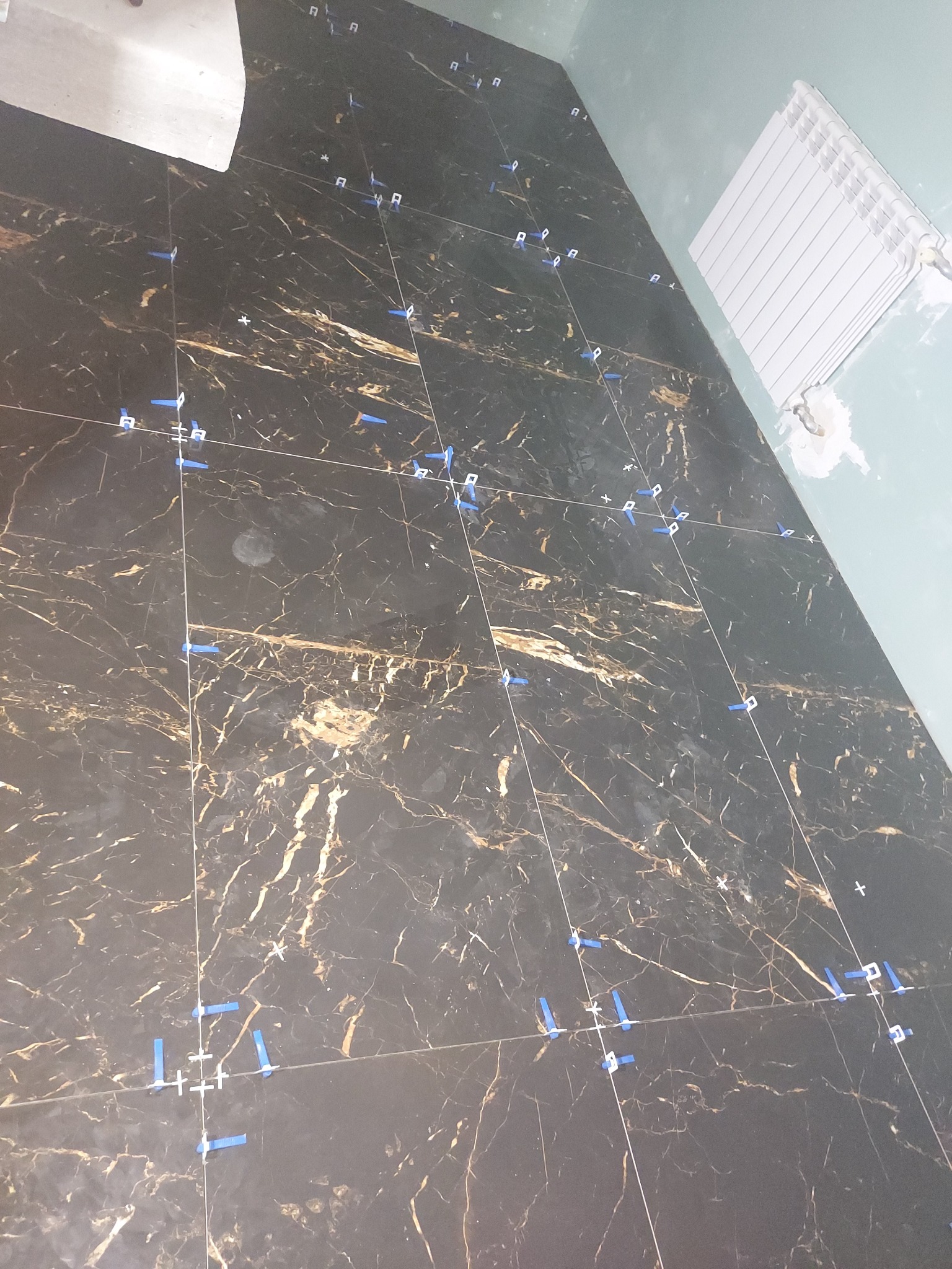 Układanie czarnych płytek podłogowych z marmurowym wzorem, z użyciem niebieskich krzyżyków dystansowych, widoczny fragment grzejnika na ścianie.