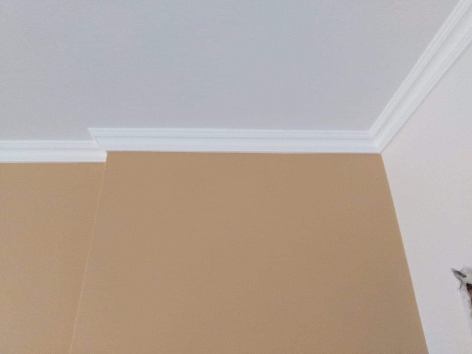 Beżowa ściana połączona z białym sufitem wykończona białą listwą dekoracyjną przy suficie.