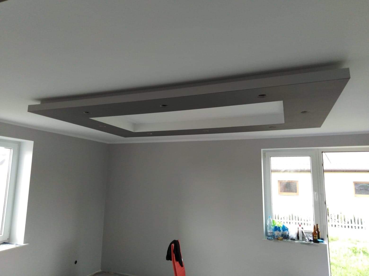 Pomieszczenie po remoncie z szarymi ścianami i podwieszanym sufitem z oświetleniem punktowym. Widoczne dwa okna i krzesło z ubraniem.