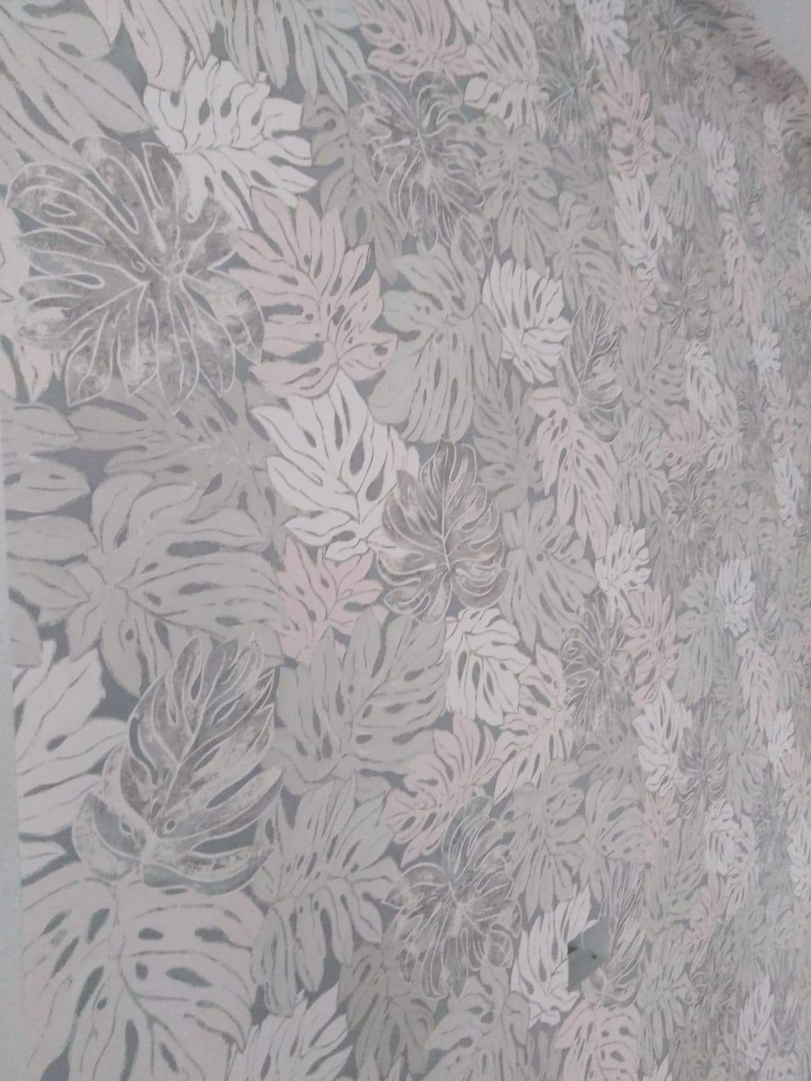 Ściana pokryta tapetą w delikatny wzór liści monstery w odcieniach szarości, bieli i pudrowego różu, widoczne z bliska pod kątem.