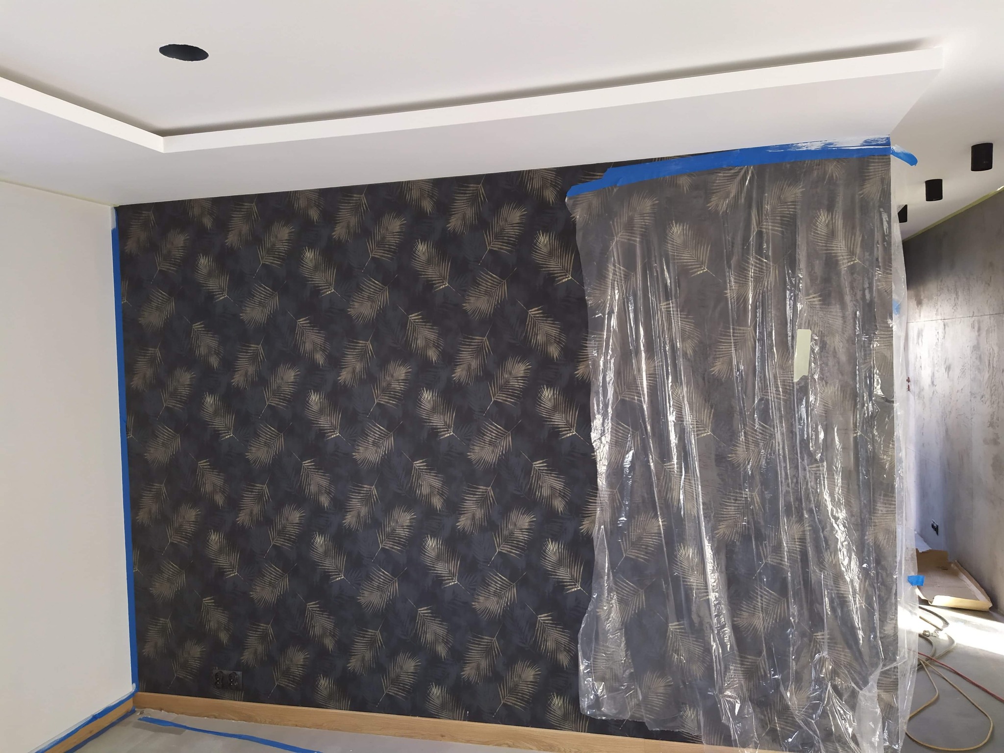 Ściana z tapetą w liście palmowe, zabezpieczona folią malarską, widoczne taśmy malarskie na podłodze i suficie, przygotowanie do prac wykończeniowych.