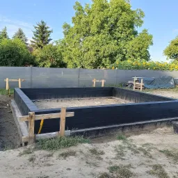 Wylany fundament z czarną izolacją przeciwwilgociową, drewniane szalunki, widok na ogród z kwitnącymi słonecznikami w tle.