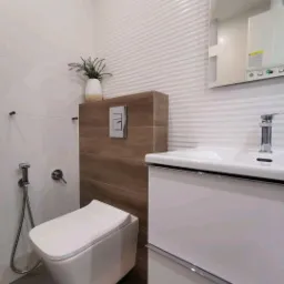 Nowoczesna łazienka z podwieszaną toaletą, bidetką i umywalką z szafką, białe płytki z falistą strukturą, drewnopodobna zabudowa stelaża toalety, dekoracyjna roślina w wazonie.