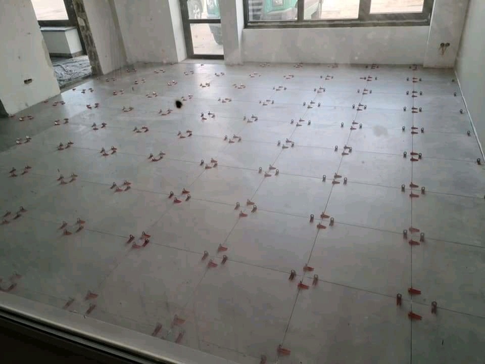 Układanie jasnych, kwadratowych płytek podłogowych z użyciem czerwonych klipsów poziomujących, widok z góry na surowy stan wnętrza.