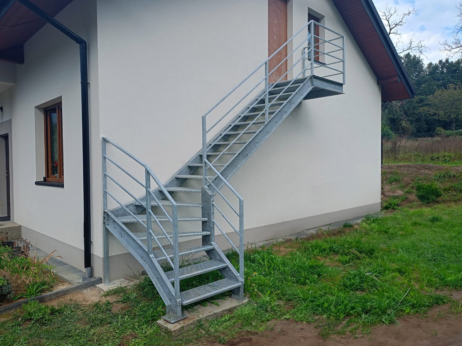 Zewnętrzne schody metalowe z poręczami prowadzące do drzwi na piętrze budynku. Schody ocynkowane, z ażurowymi stopniami. Widok z dołu, zieleń w tle.