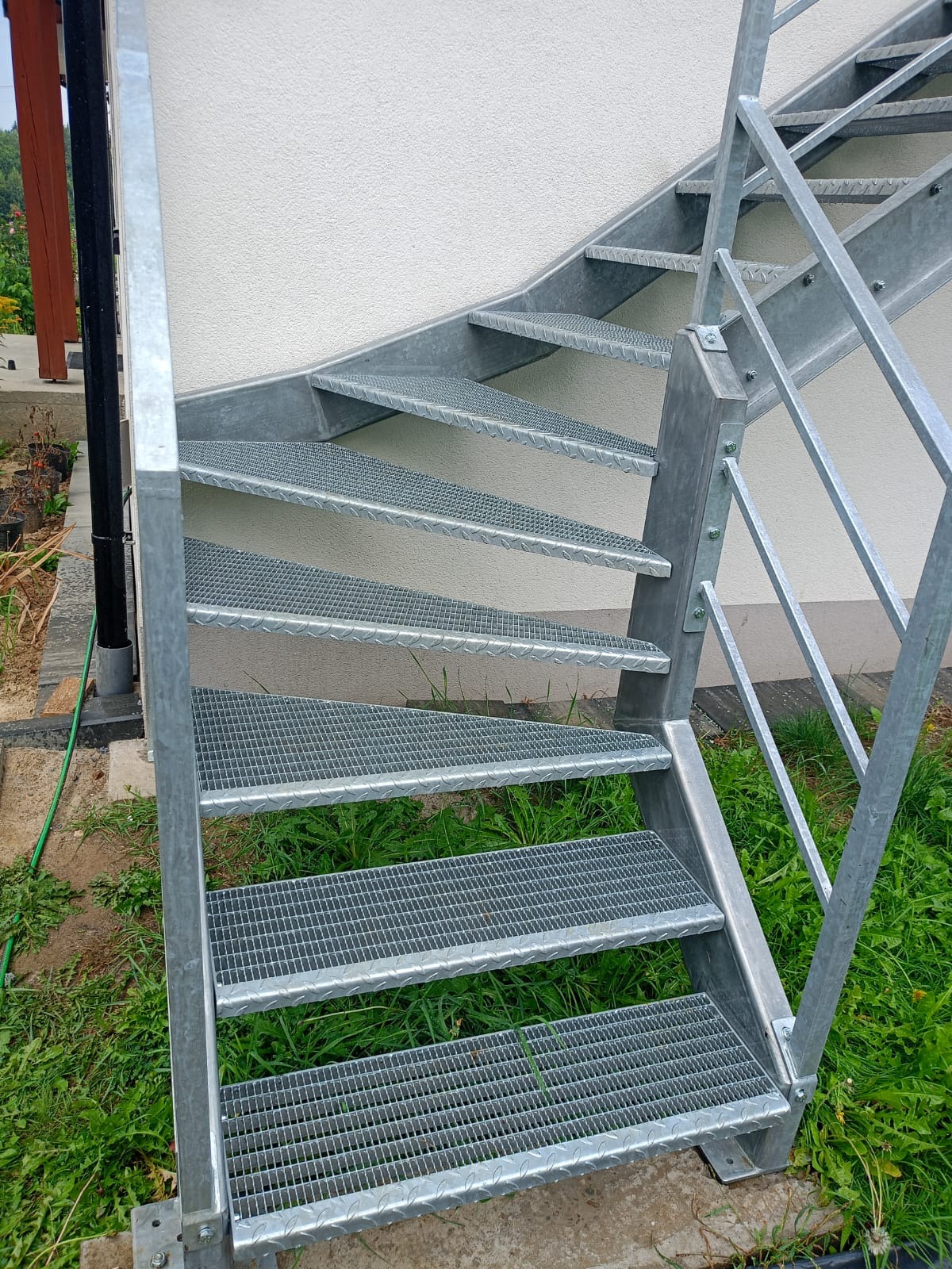 Zewnętrzne schody metalowe z ażurowymi stopniami i prostą balustradą, prowadzące wzdłuż jasnej ściany budynku, otoczone zielenią. Widok z dołu, podkreślający konstrukcję.