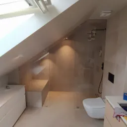 Jasna łazienka na poddaszu ze skośnym sufitem, kabiną prysznicową ze szklanymi ściankami, podwieszaną toaletą i zabudowaną umywalką z szafkami.