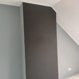 Ściana w pokoju na poddaszu, pomalowana na szaro-niebieski kolor z pionowym pasem w kolorze grafitowym, widoczna listwa przysufitowa i gniazdko elektryczne.