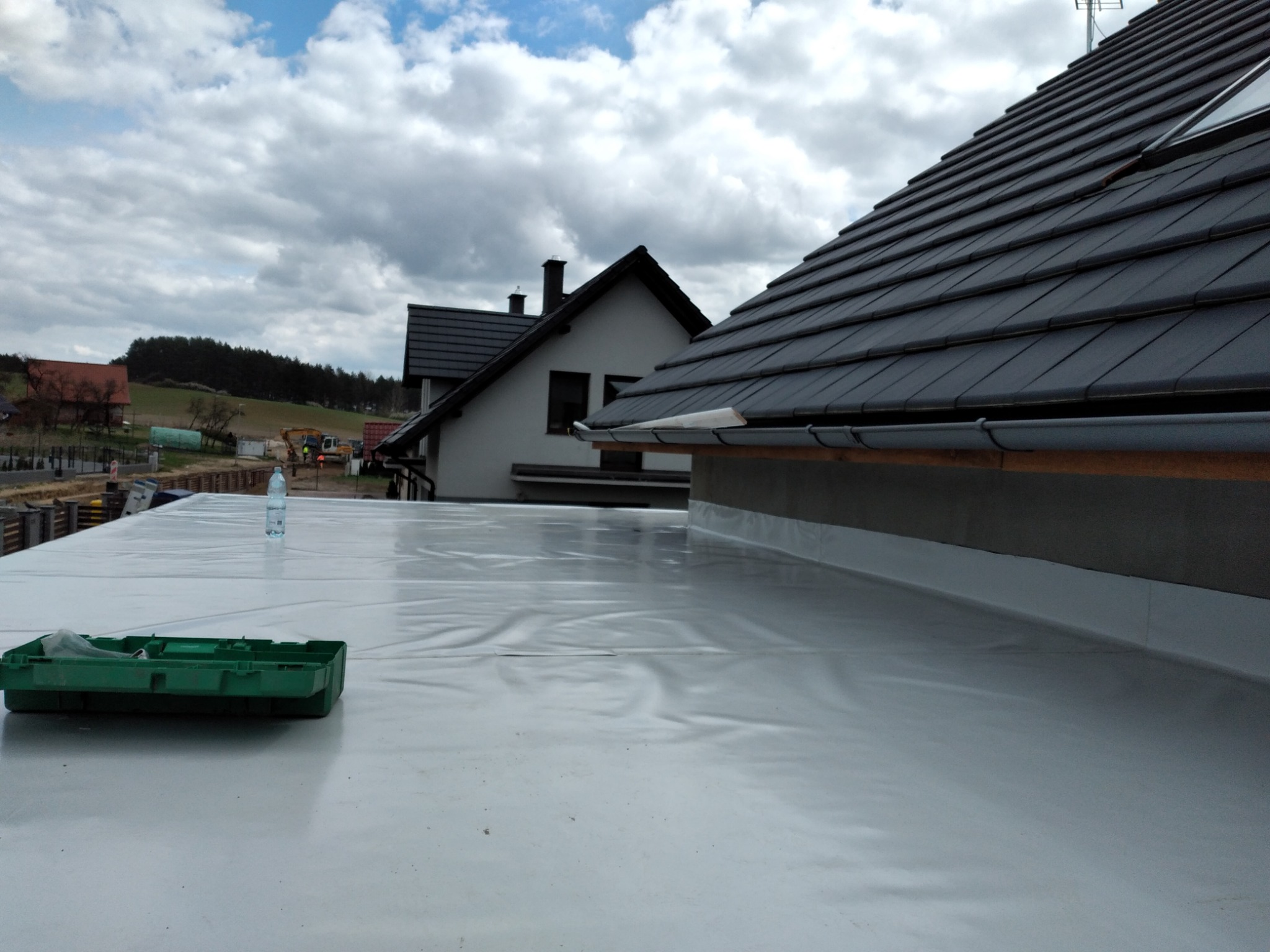 Biała membrana dachowa rozłożona na płaskim dachu, z widocznym zielonym pojemnikiem na narzędzia i butelką wody w tle, fragment dachu z szarą dachówką i elementami orynnowania, w tle widoczne...