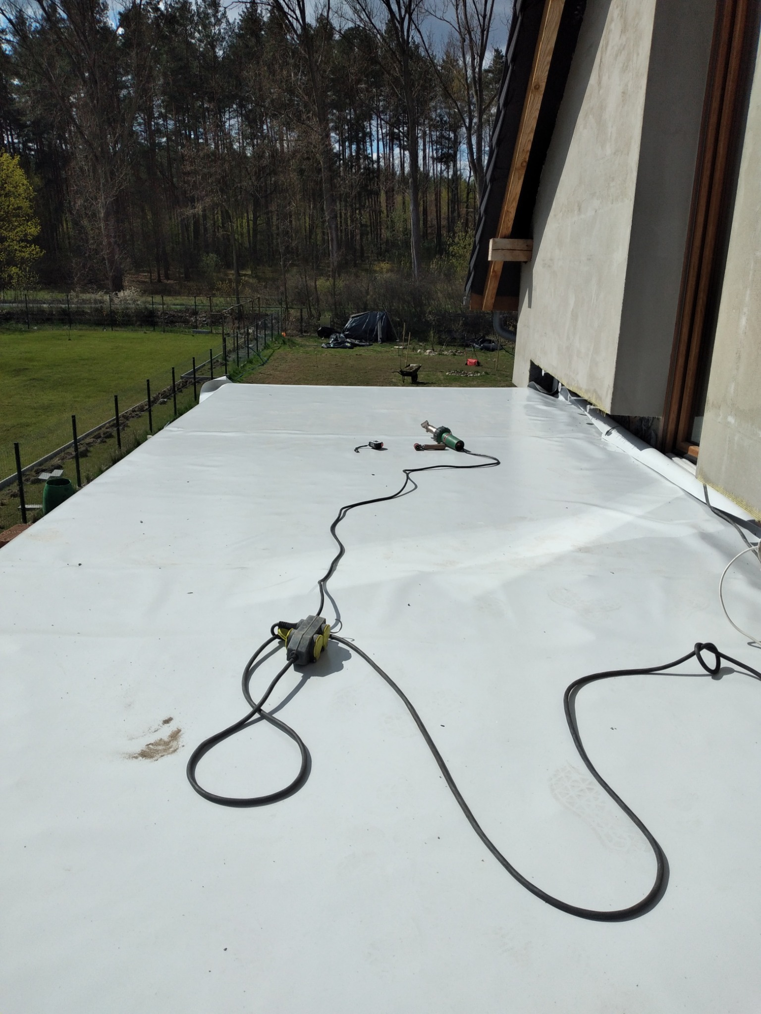 Biała membrana dachowa rozłożona na płaskim dachu, widoczny sprzęt do zgrzewania: zgrzewarka i przedłużacz, w tle budynek z drewnianą konstrukcją dachu i zielony krajobraz z drzewami.