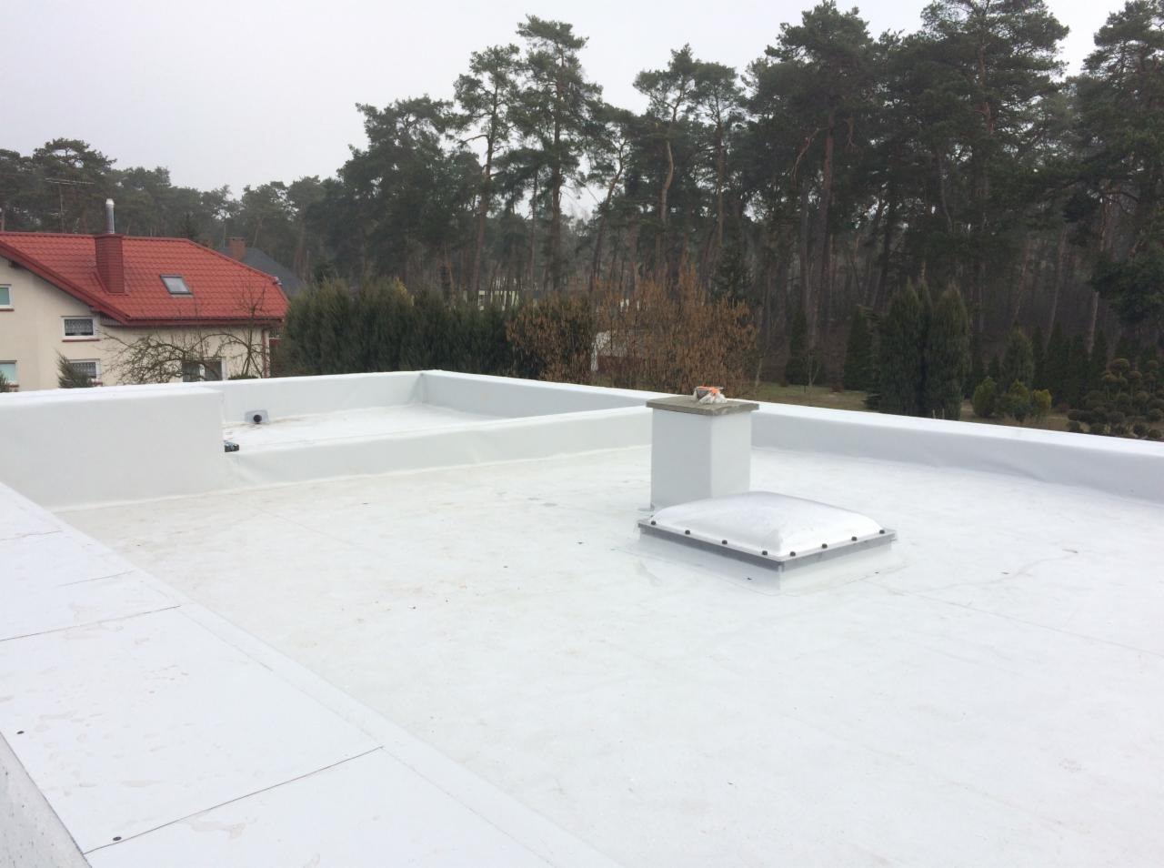 Biały, płaski dach pokryty membraną, z kominem i świetlikiem, na tle drzew i zabudowań.