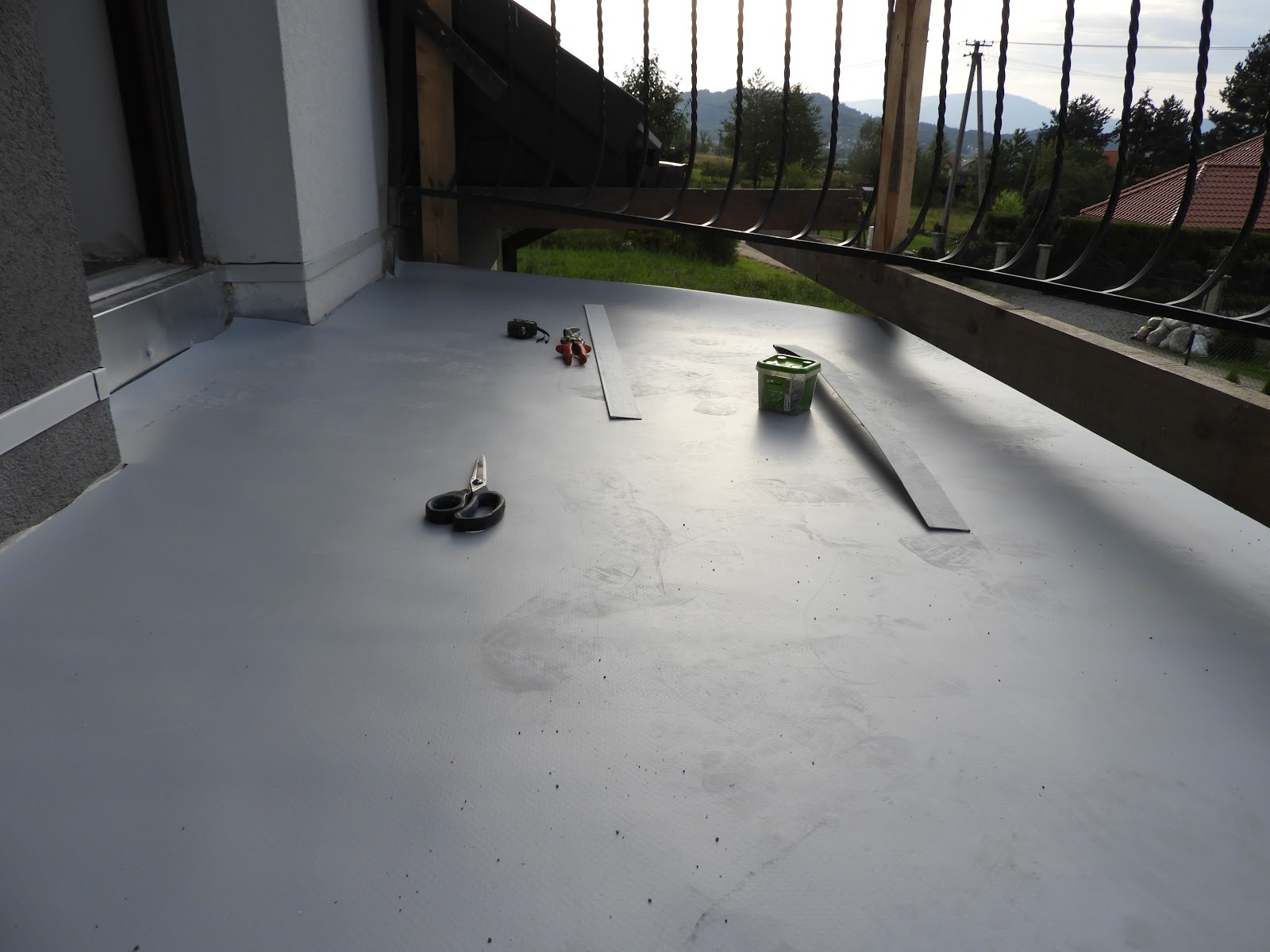 Szara, płaska powierzchnia dachu z widocznymi narzędziami dekarskimi: nożyczki, miara, puszka z klejem i metalowe listwy, na tle balustrady i krajobrazu.