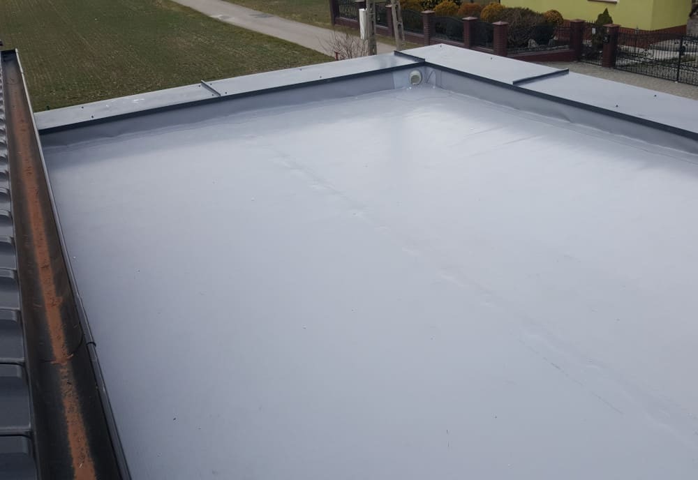 Szary, płaski dach pokryty membraną PVC z widocznym kominkiem wentylacyjnym i fragmentem dachu sąsiedniego budynku z dachówką ceramiczną w tle.