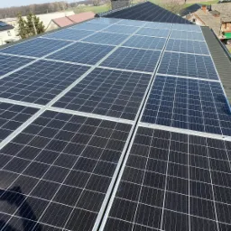 Rząd paneli fotowoltaicznych na dachu budynku gospodarczego z widocznymi zabudowaniami w tle, wykonany w słoneczny dzień.