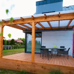 Nowoczesny taras z drewna z zadaszeniem z przezroczystych paneli, wyposażony w stół i krzesła z rattanu, przylegający do domu z elementami w kolorze szarym i czerwonym, w tle widoczna altana.